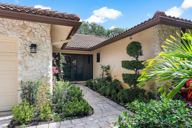 entrance to property featuring a garage, stone siding, a tile roof, and stucco siding