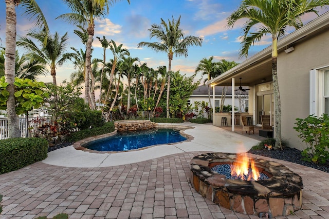 pool at dusk with ceiling fan, a patio, exterior kitchen, and a fire pit