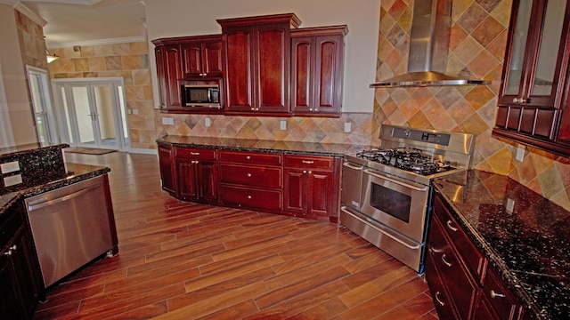 kitchen with wall chimney range hood, appliances with stainless steel finishes, backsplash, ornamental molding, and dark stone counters