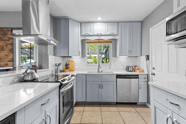 kitchen featuring appliances with stainless steel finishes, light stone countertops, island exhaust hood, gray cabinets, and sink