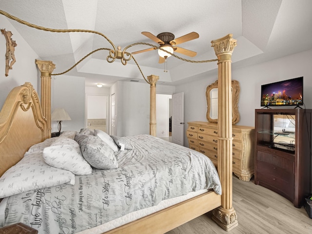 bedroom with ornate columns, ceiling fan, a tray ceiling, ensuite bath, and light hardwood / wood-style flooring