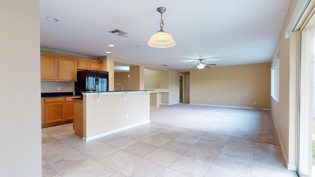 kitchen with ceiling fan, hanging light fixtures, a center island with sink, light carpet, and black fridge