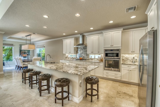 kitchen featuring pendant lighting, appliances with stainless steel finishes, an island with sink, a kitchen bar, and wall chimney exhaust hood