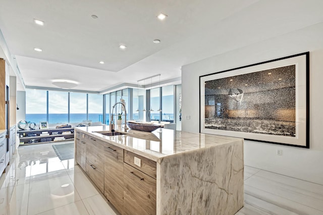 kitchen with sink, floor to ceiling windows, a water view, light stone countertops, and an island with sink