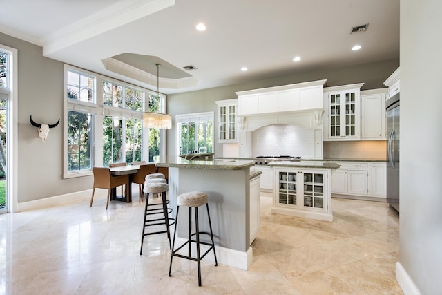 kitchen with a center island with sink, pendant lighting, a raised ceiling, crown molding, and white cabinetry