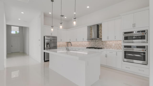 kitchen featuring wall chimney exhaust hood, white cabinetry, decorative light fixtures, appliances with stainless steel finishes, and an island with sink