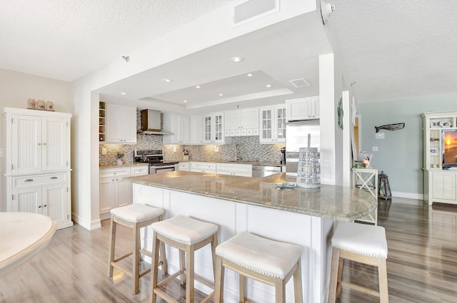 kitchen featuring appliances with stainless steel finishes, a breakfast bar, white cabinetry, a raised ceiling, and wall chimney exhaust hood