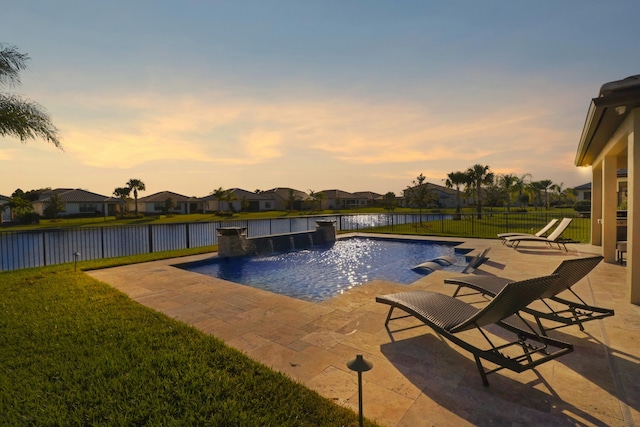pool at dusk with a patio, a water view, a yard, and pool water feature