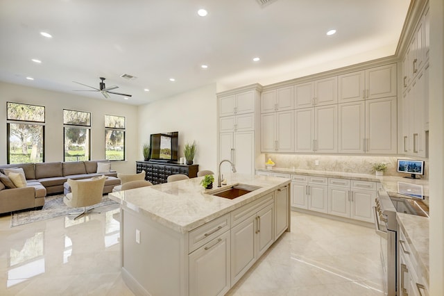 kitchen with sink, light stone counters, tasteful backsplash, stainless steel stove, and a kitchen island with sink