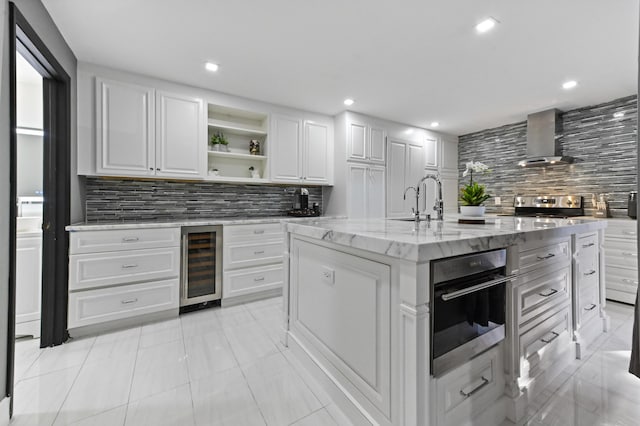 kitchen with white cabinets, decorative backsplash, light stone countertops, a center island with sink, and wall chimney exhaust hood