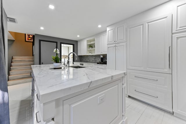 kitchen featuring sink, a kitchen island with sink, white cabinetry, light stone counters, and decorative backsplash