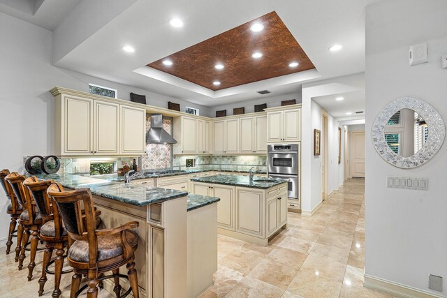 kitchen featuring sink, wall chimney exhaust hood, a center island with sink, cream cabinetry, and dark stone counters