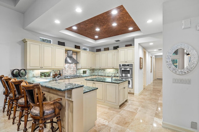 kitchen with cream cabinetry, a raised ceiling, kitchen peninsula, and wall chimney exhaust hood