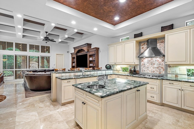 kitchen with sink, dark stone countertops, a center island with sink, cream cabinetry, and wall chimney exhaust hood