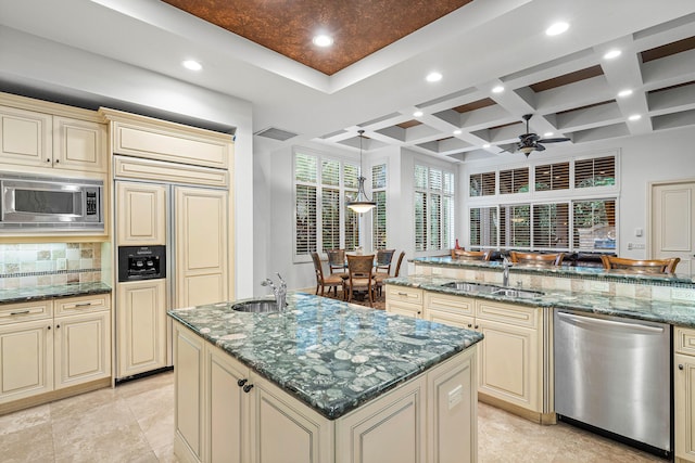 kitchen with an island with sink, sink, dark stone counters, built in appliances, and cream cabinetry