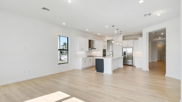 kitchen featuring stainless steel fridge, white cabinetry, hanging light fixtures, an island with sink, and wall chimney exhaust hood