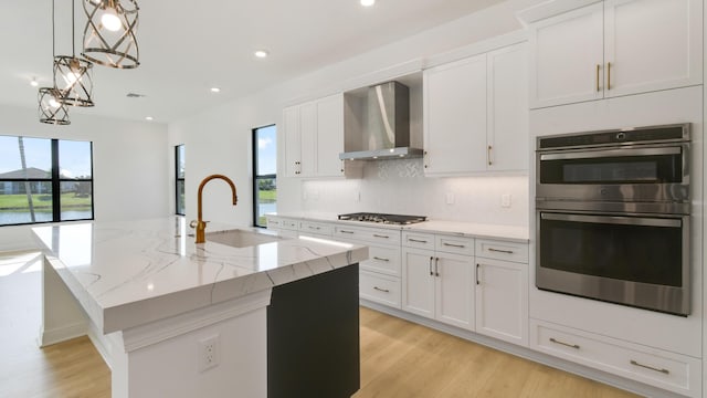 kitchen with sink, white cabinetry, appliances with stainless steel finishes, an island with sink, and wall chimney range hood