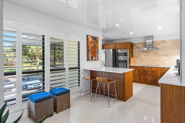 kitchen featuring wall chimney range hood, appliances with stainless steel finishes, tasteful backsplash, a kitchen bar, and light tile patterned flooring