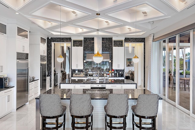 kitchen with a kitchen bar, stainless steel built in refrigerator, coffered ceiling, wall chimney exhaust hood, and a large island with sink