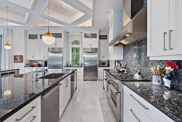 kitchen featuring premium appliances, a sink, glass insert cabinets, wall chimney range hood, and tasteful backsplash