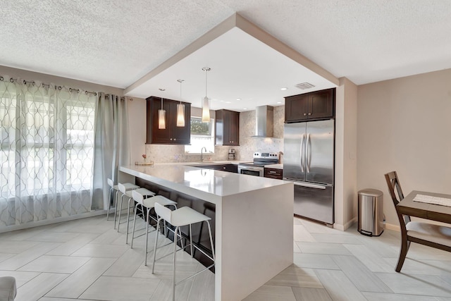 kitchen featuring a kitchen bar, decorative light fixtures, appliances with stainless steel finishes, kitchen peninsula, and wall chimney range hood