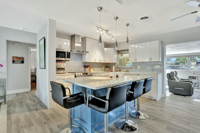 kitchen featuring wall chimney range hood, a breakfast bar area, appliances with stainless steel finishes, white cabinetry, and decorative light fixtures