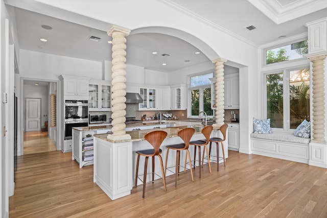 kitchen with ornate columns, white cabinetry, a center island with sink, and light stone counters