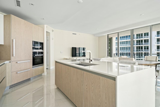 kitchen featuring black double oven, an island with sink, sink, and light brown cabinetry