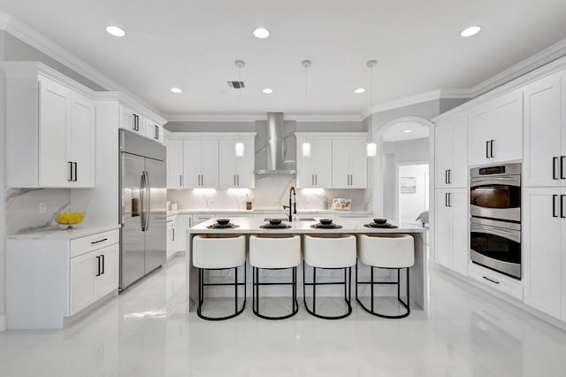 kitchen with white cabinetry, decorative light fixtures, appliances with stainless steel finishes, a kitchen bar, and wall chimney exhaust hood