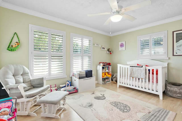 bedroom featuring a nursery area, light wood-type flooring, ornamental molding, ceiling fan, and a textured ceiling