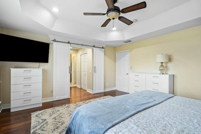 bedroom with a barn door, dark hardwood / wood-style floors, a tray ceiling, and ceiling fan