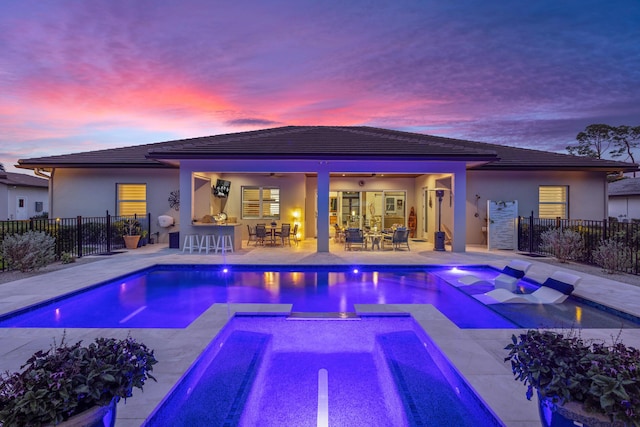 pool at dusk with a fenced in pool, outdoor dry bar, a ceiling fan, fence, and an in ground hot tub