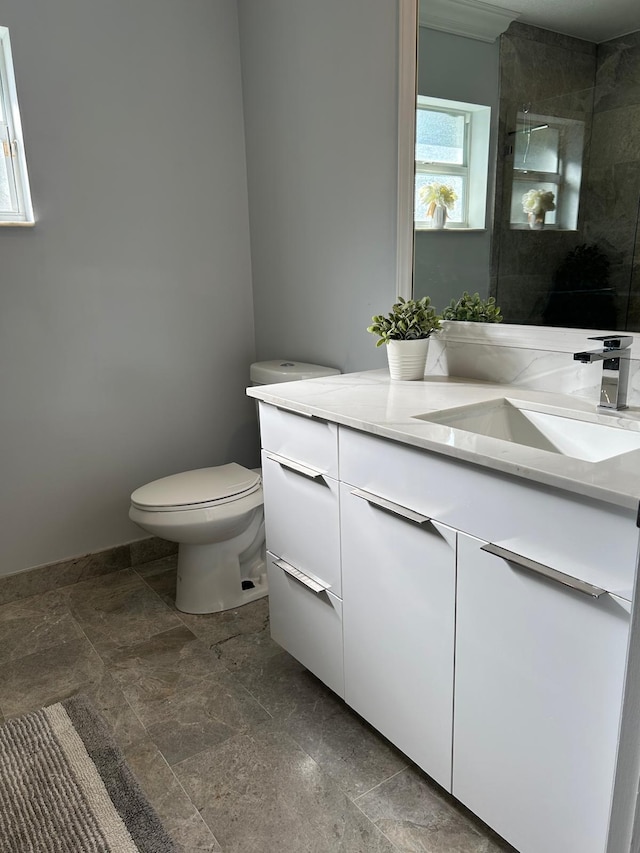 full bathroom featuring toilet, vanity, tiled shower, stone finish floors, and baseboards