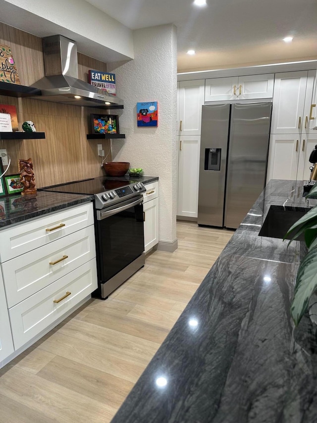 kitchen with appliances with stainless steel finishes, white cabinetry, light hardwood / wood-style floors, dark stone counters, and wall chimney exhaust hood