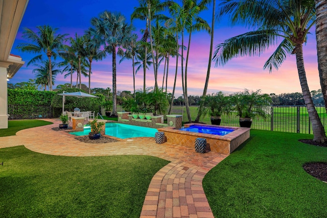 pool at dusk featuring an in ground hot tub, a patio area, and a lawn