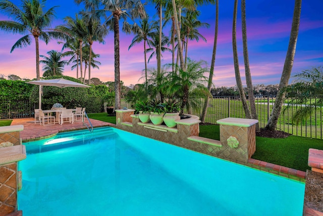 pool at dusk with a patio area and a lawn