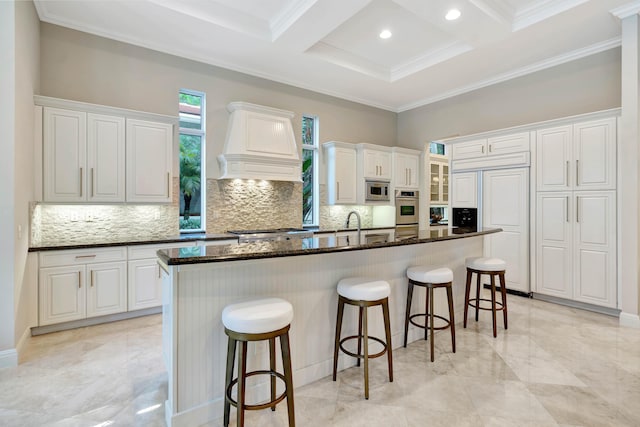 kitchen featuring premium range hood, built in appliances, dark stone countertops, a large island with sink, and white cabinets