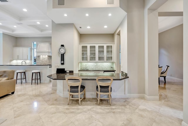 kitchen with coffered ceiling, a breakfast bar area, kitchen peninsula, dark stone counters, and white cabinets