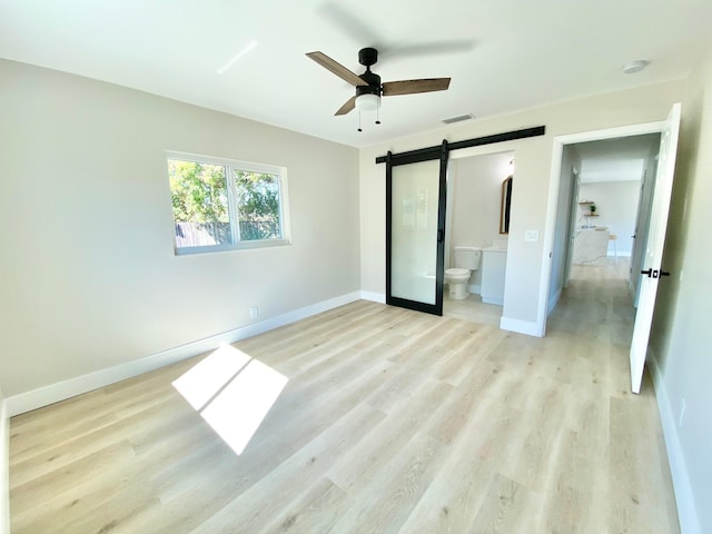 unfurnished bedroom featuring ensuite bath, a barn door, ceiling fan, and light wood-type flooring