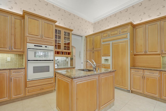 kitchen with sink, a kitchen island with sink, double oven, paneled built in fridge, and light stone countertops