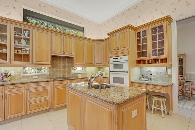 kitchen with sink, double oven, black electric stovetop, light stone countertops, and an island with sink