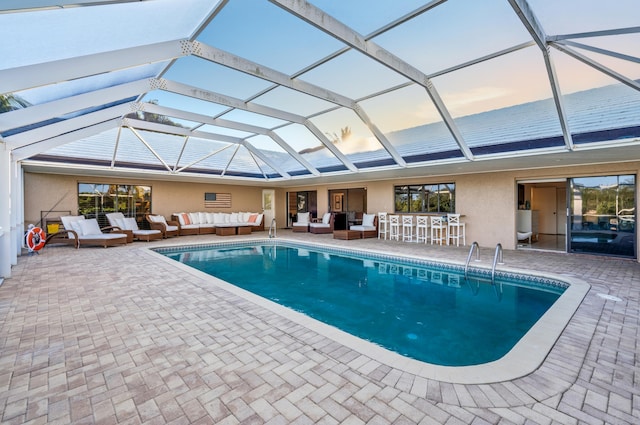 pool at dusk featuring outdoor lounge area, a patio, glass enclosure, and exterior bar