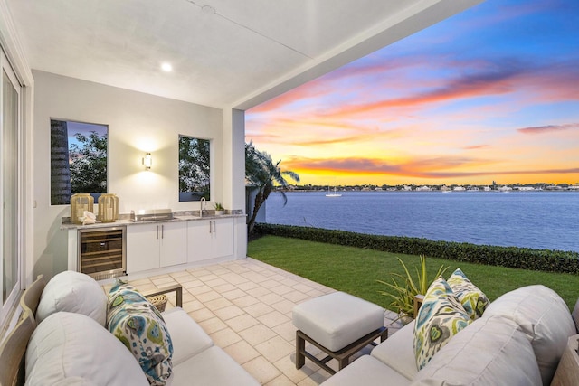 patio terrace at dusk with beverage cooler, a water view, a lawn, and area for grilling