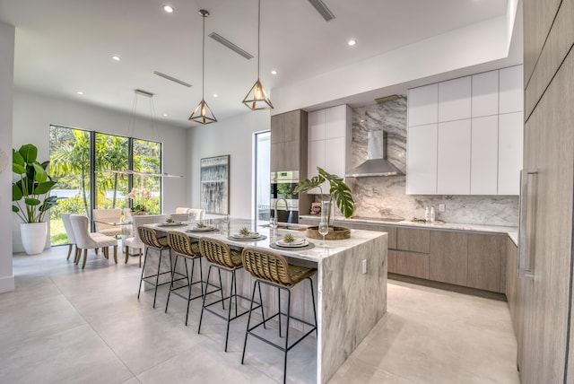 kitchen with white cabinetry, hanging light fixtures, backsplash, an island with sink, and wall chimney exhaust hood