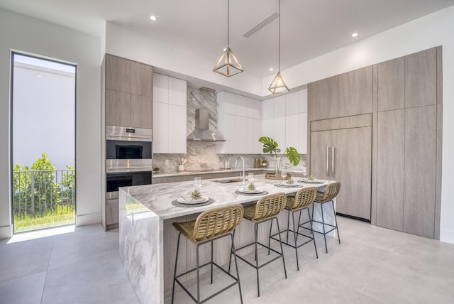 kitchen with sink, a kitchen island with sink, wall chimney exhaust hood, tasteful backsplash, and stainless steel double oven