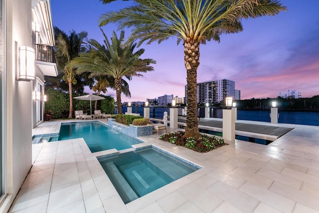 pool at dusk with a water view, a pool with connected hot tub, a patio area, and a city view