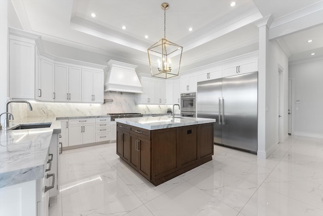 kitchen featuring custom range hood, appliances with stainless steel finishes, a center island with sink, a raised ceiling, and a sink