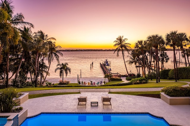 pool at dusk featuring an outdoor pool, a water view, and a patio