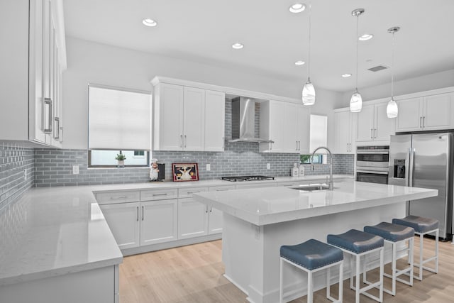 kitchen with sink, white cabinets, wall chimney range hood, stainless steel appliances, and a center island with sink