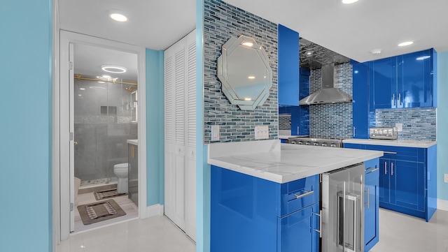 kitchen with wine cooler, wall chimney range hood, light stone counters, and blue cabinetry